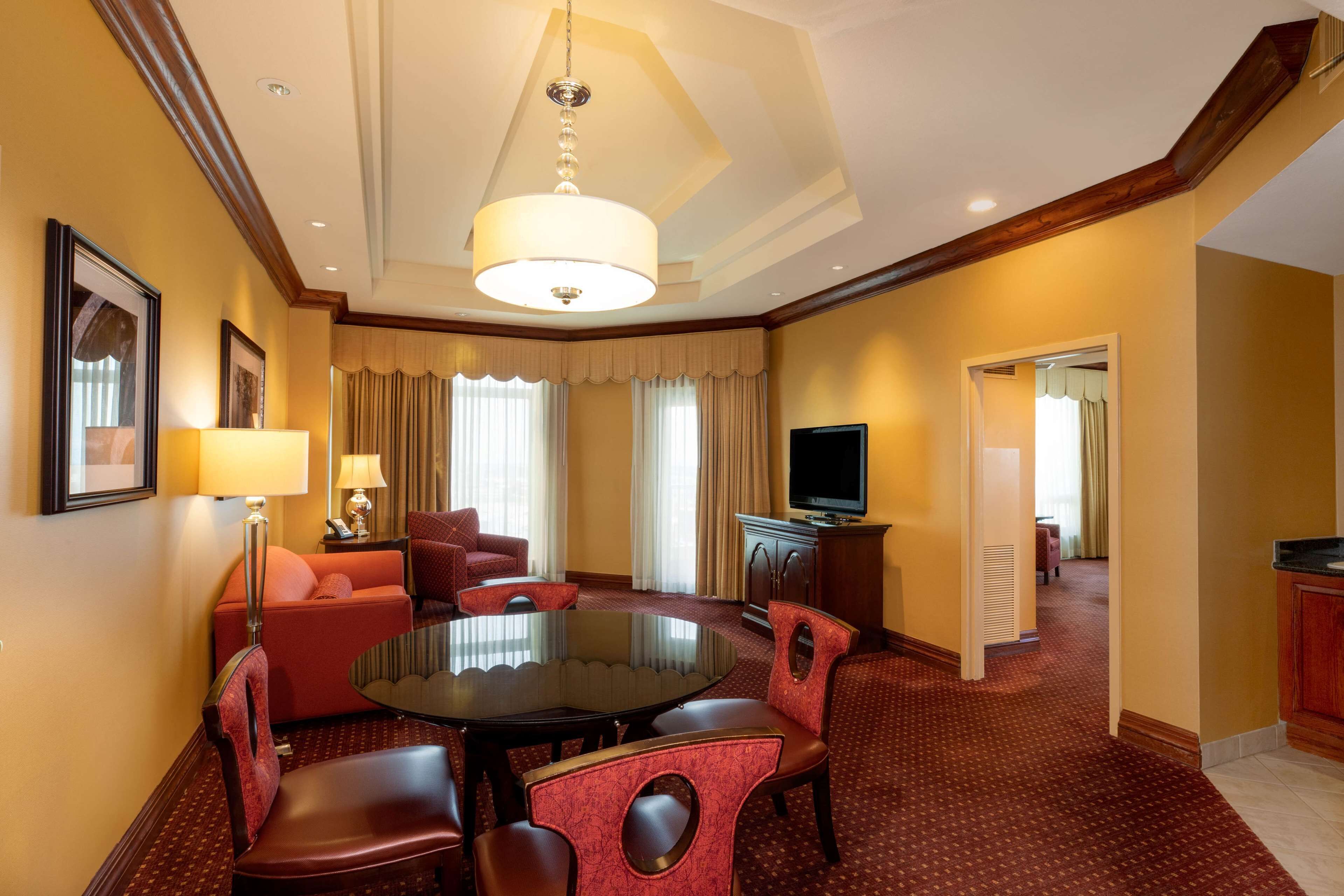 A hotel room with a dining area featuring a round table and red upholstered chairs, a sofa, a television, and a large window with curtains.