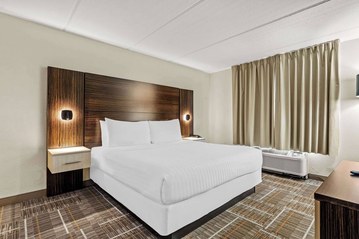 Une chambre d’hôtel avec un grand lit, une table de chevet, une lampe fixée au mur, un appareil de climatisation et une fenêtre avec des rideaux.