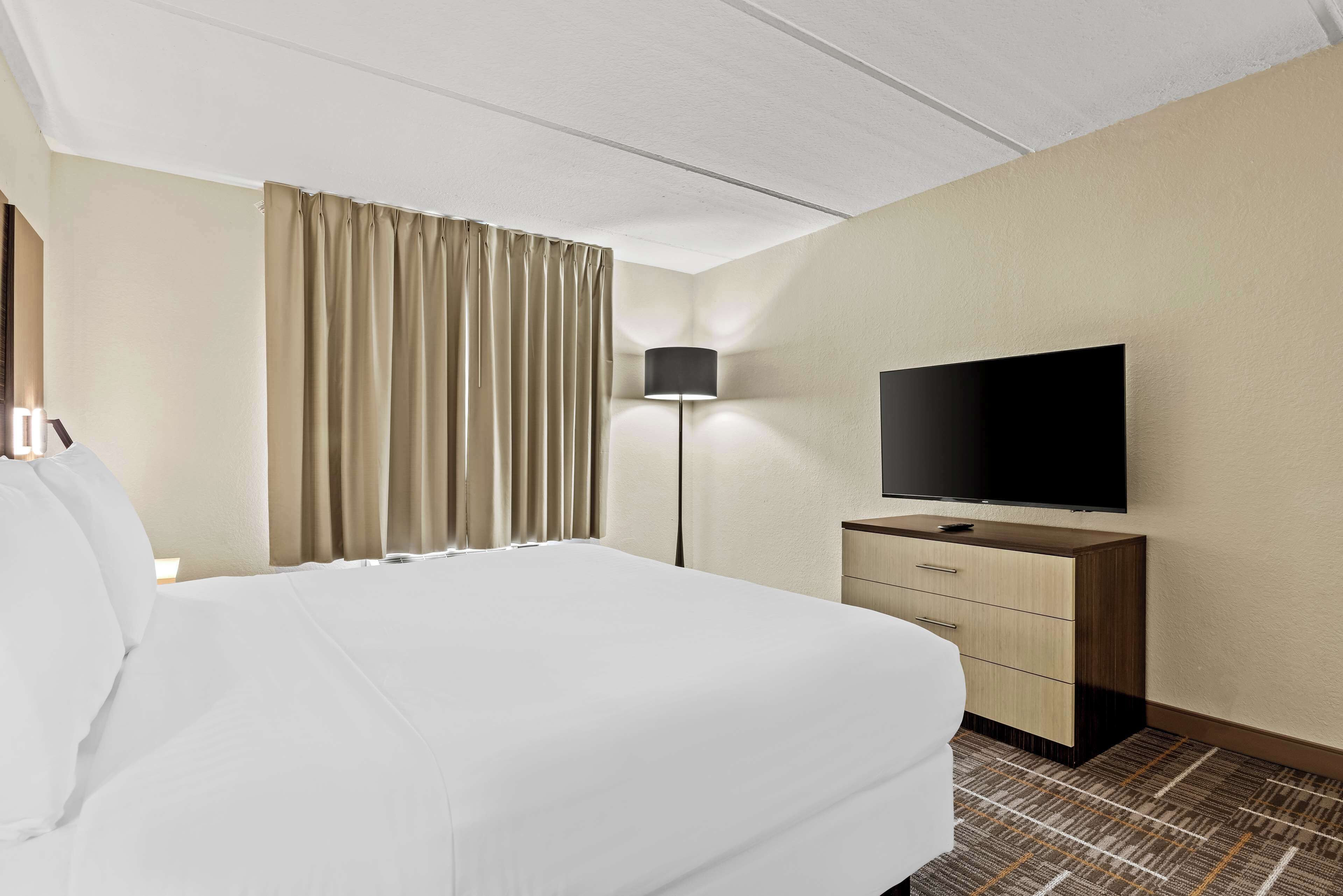 Hotelkamer met een groot bed, een flatscreen televisie op een houten dressoir, een lamp en een raam met gordijnen.