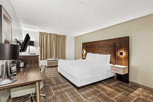 A hotel room with a large bed, a desk with a flat-screen TV, a lamp, and a nightstand with a drawer.
