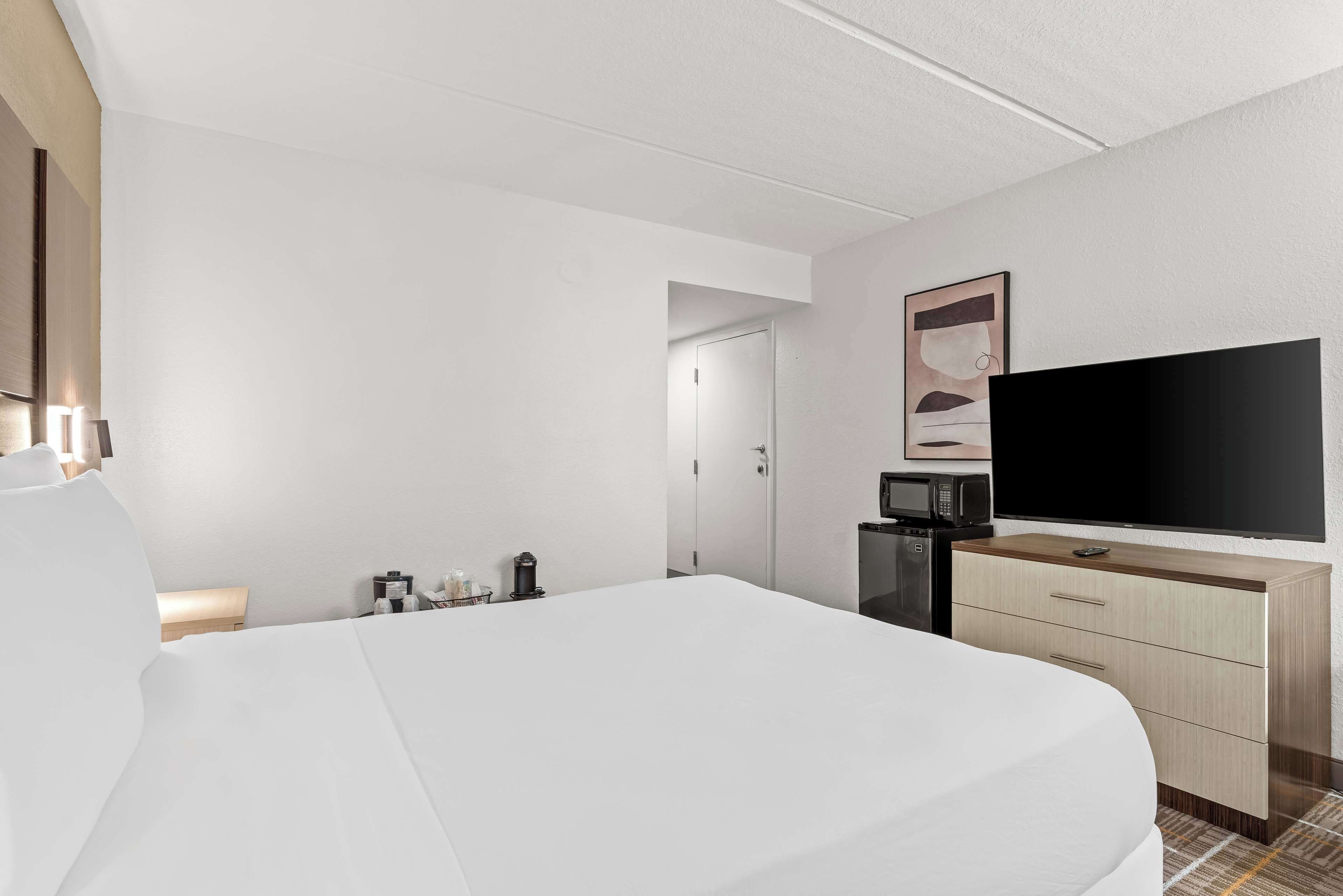 A hotel room with a bed, a flat-screen TV, a wooden dresser, a microwave, and a door leading to another room.