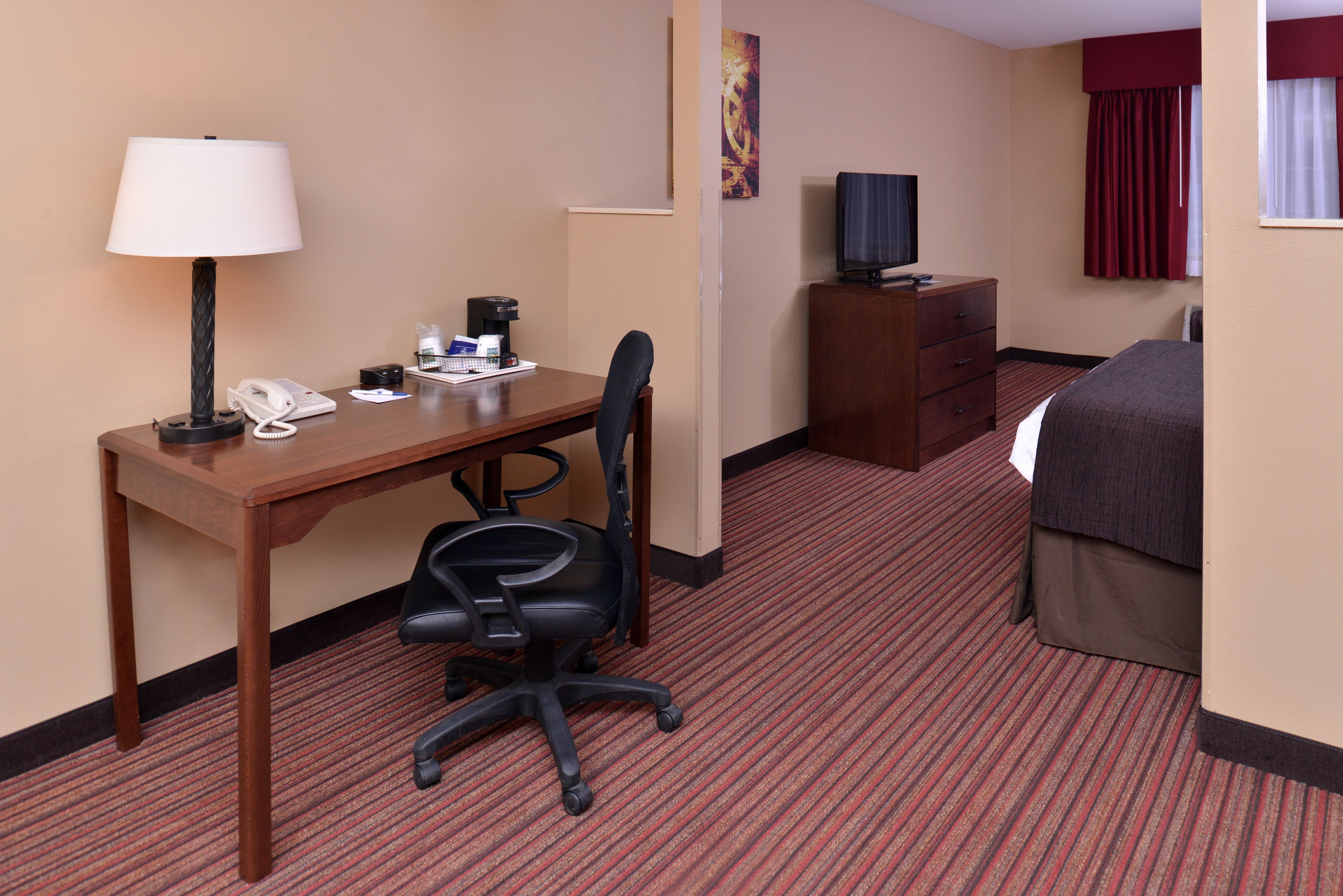 A hotel room with a wooden desk, a black office chair, a bed, a TV, a dresser, and a red carpeted floor.