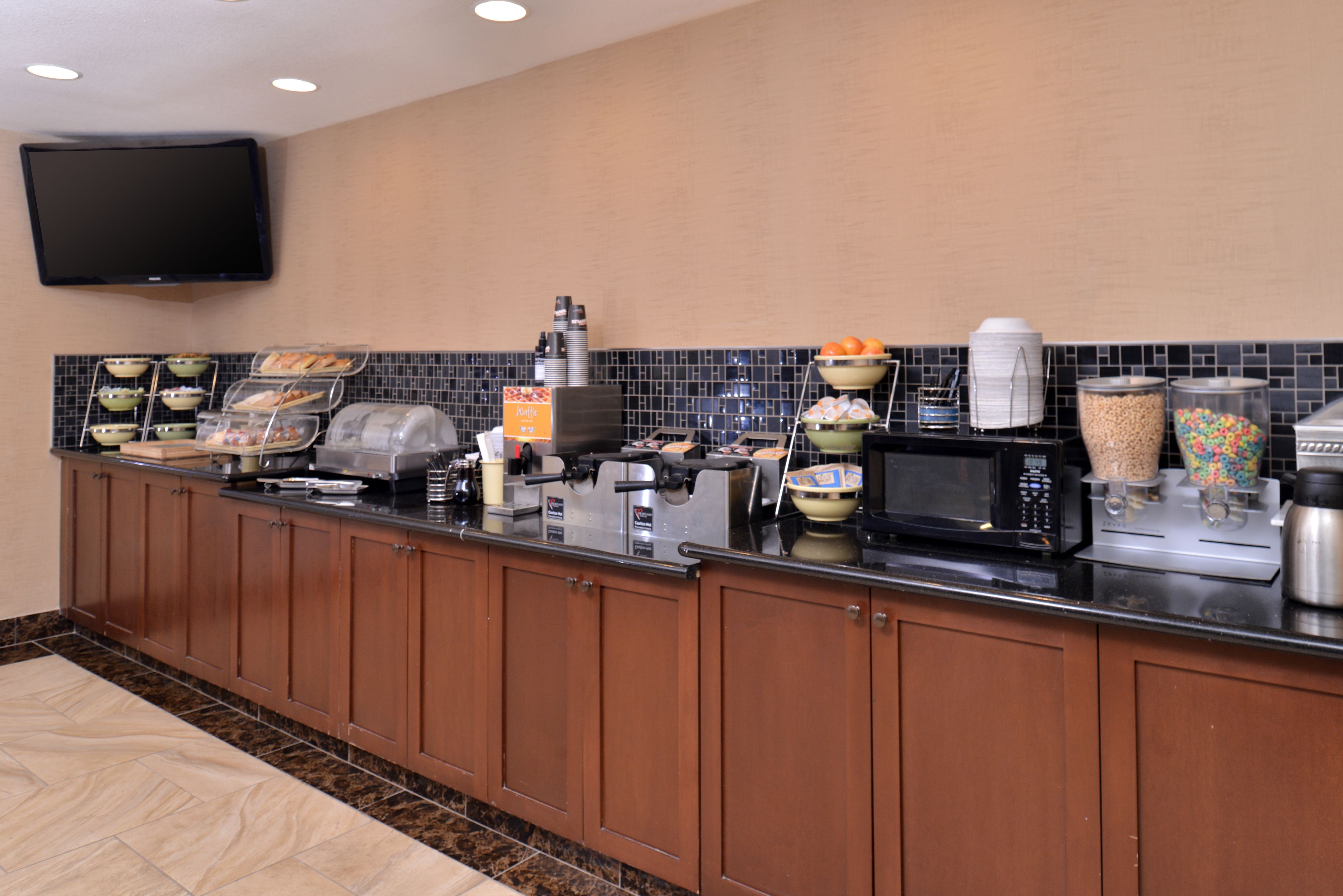 Una cocina de hotel con barra de desayuno tipo autoservicio, microondas, cafetera y varios recipientes de comida.