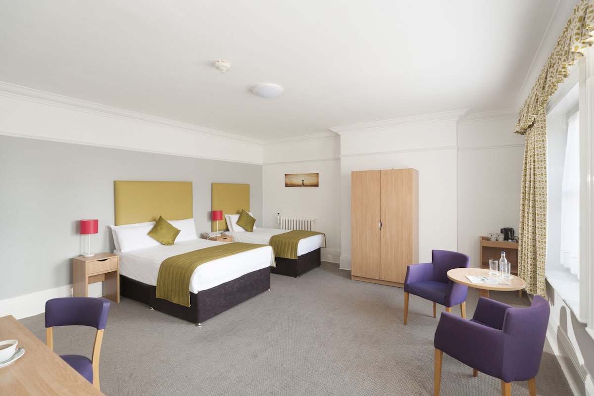 Quarto de hotel com duas camas, um guarda-roupa, uma mesa redonda, duas cadeiras roxas, uma mesinha de apoio com uma luminária e uma janela com cortinas.