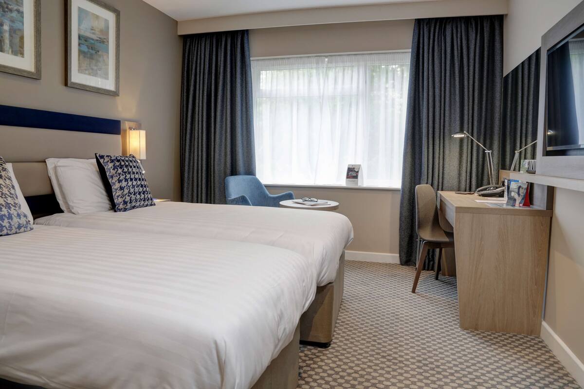 Ein Hotelzimmer mit einem großen Bett, einem Schreibtisch, einem Stuhl und einem Fenster mit Vorhängen.