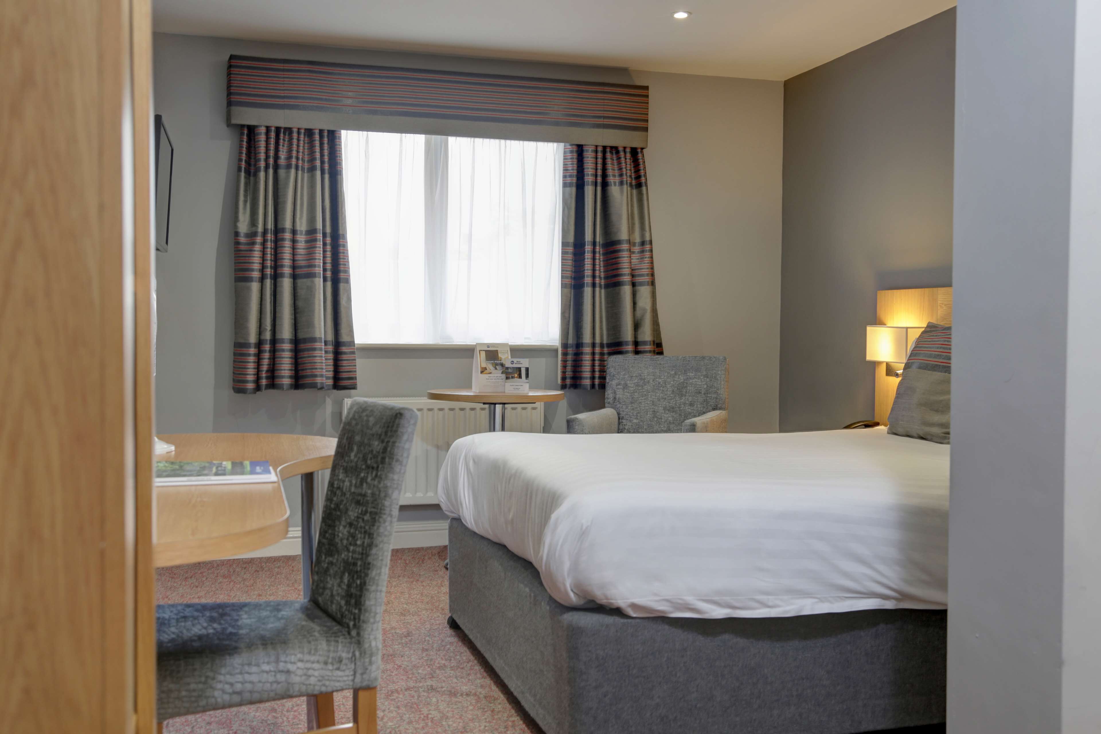 A hotel room with a bed, a desk, a chair, a lamp, and a window with curtains.