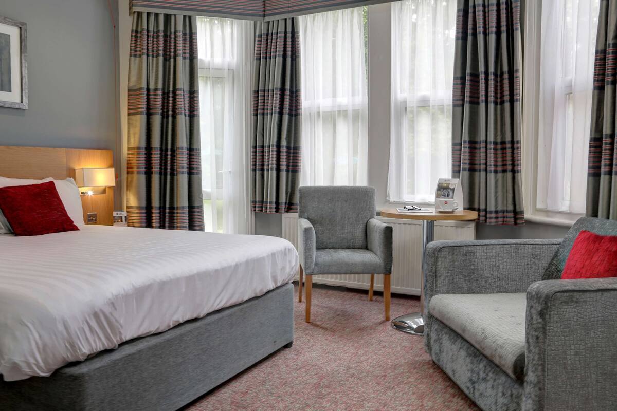 Ein Hotelzimmer mit einem Bett, zwei Sesseln, einem kleinen Tisch mit Kaffeemaschine und einem Fenster mit Vorhängen.
