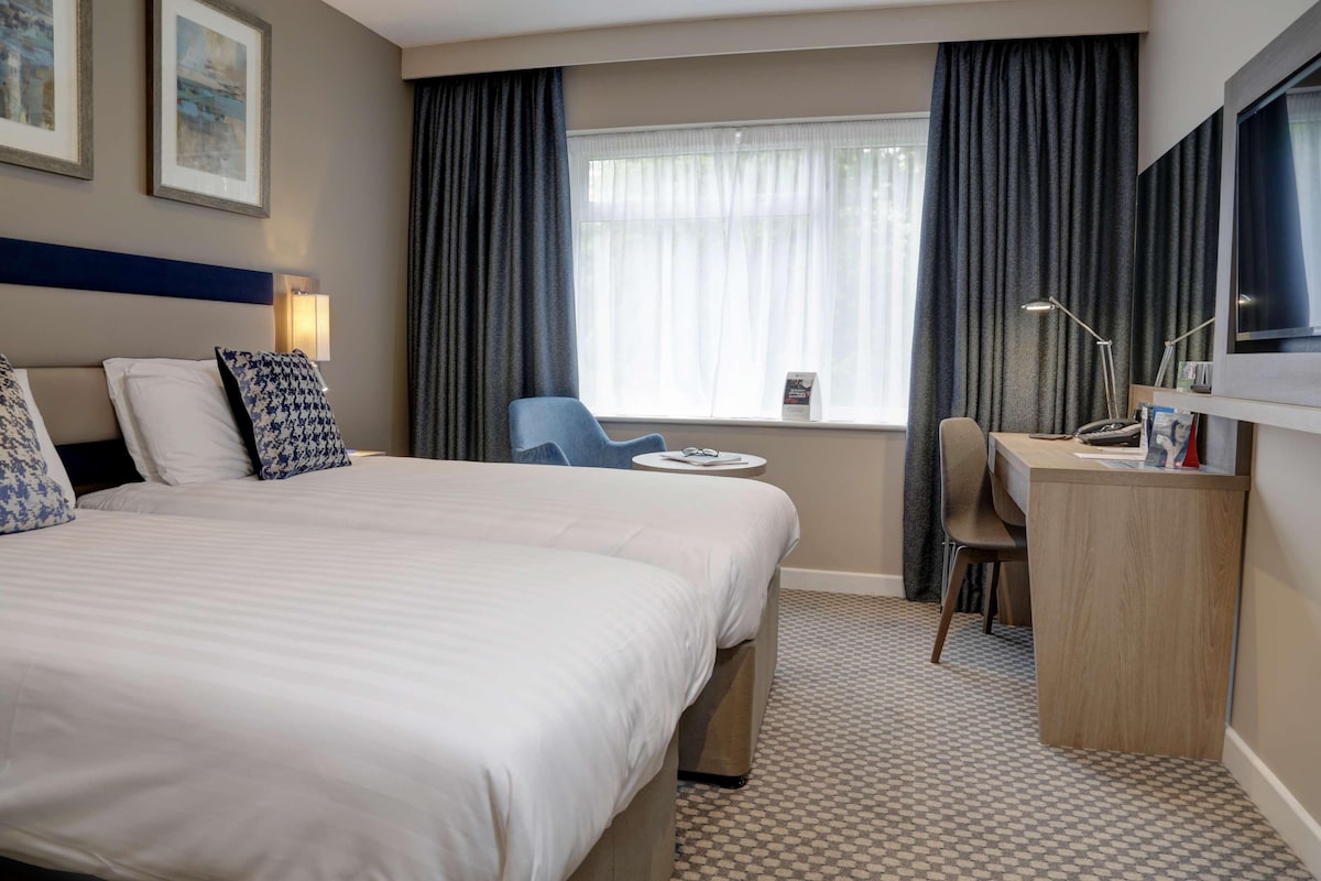 Une chambre d’hôtel avec un grand lit, un bureau, une chaise, une fenêtre avec des rideaux et des œuvres d’art encadrées accrochées au mur.