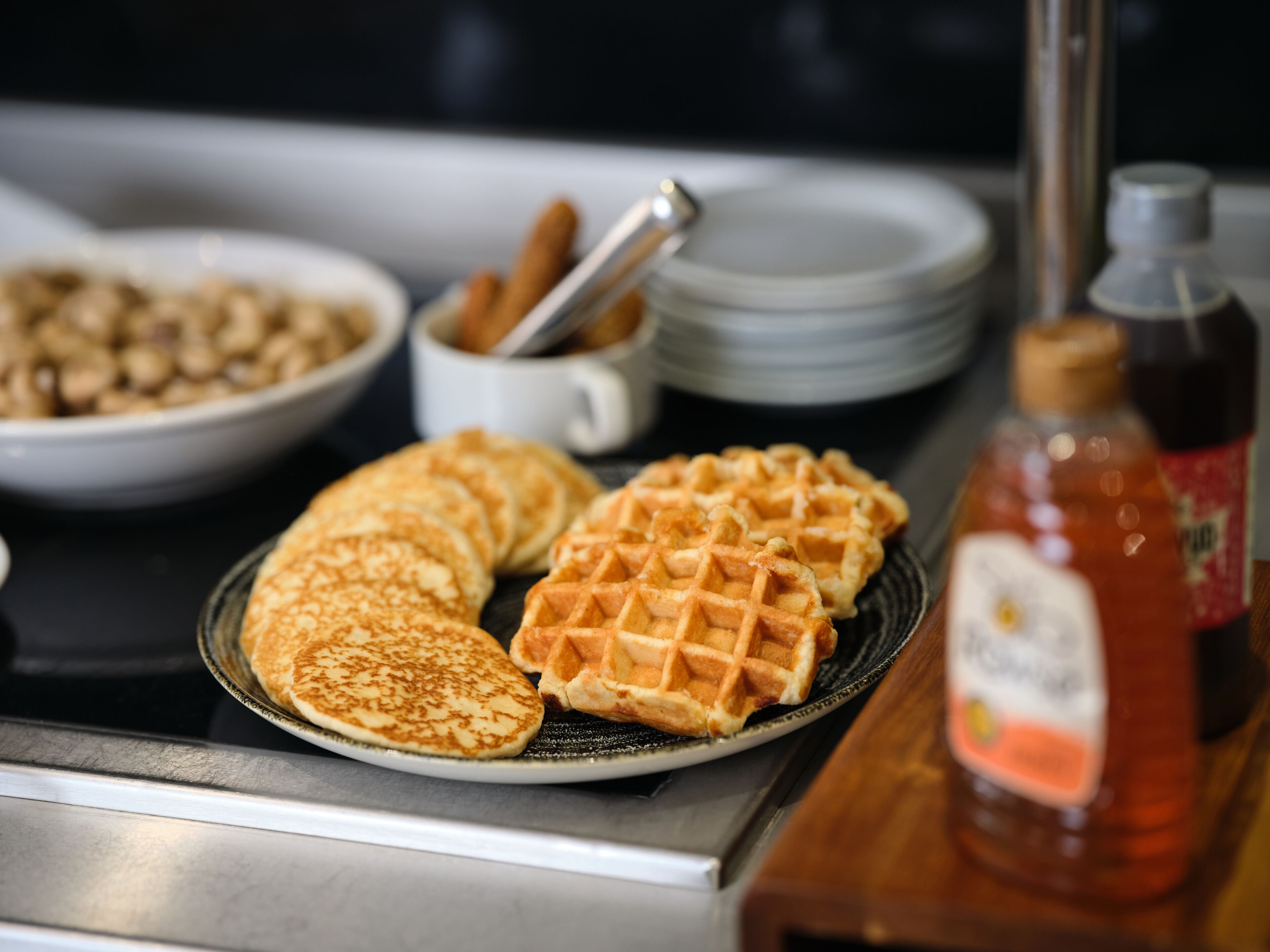 A breakfast setting with pancakes and waffles on a plate, a bowl of cereal, a cup with a spoon, and a bottle of syrup.