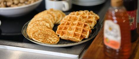 A breakfast setting with pancakes and waffles on a plate, a bowl of cereal, a cup with a spoon, and a bottle of syrup.