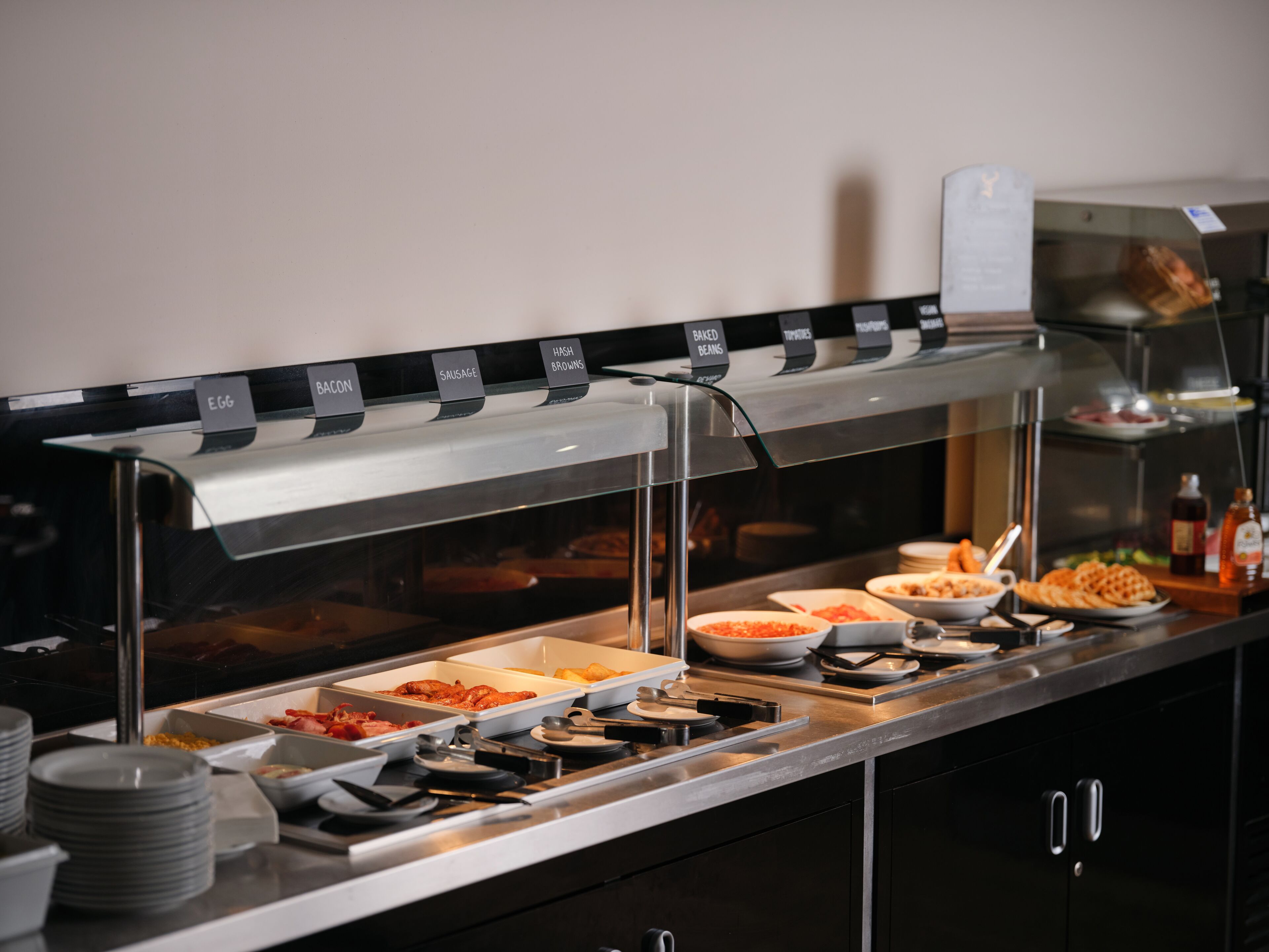Un buffet proposant différents plats, chacun clairement identifié, notamment des œufs, du bacon, des saucisses et des haricots cuits.