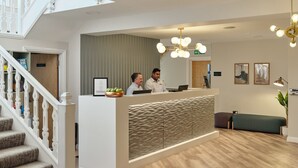 Un espace d’accueil comprenant un comptoir de réception, deux employés, un escalier, un banc vert et un luminaire au plafond.