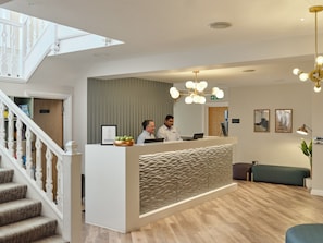 Ein Empfangsbereich mit einer Rezeption, zwei Mitarbeitern, einer Treppe, einer grünen Bank und einer Deckenleuchte.