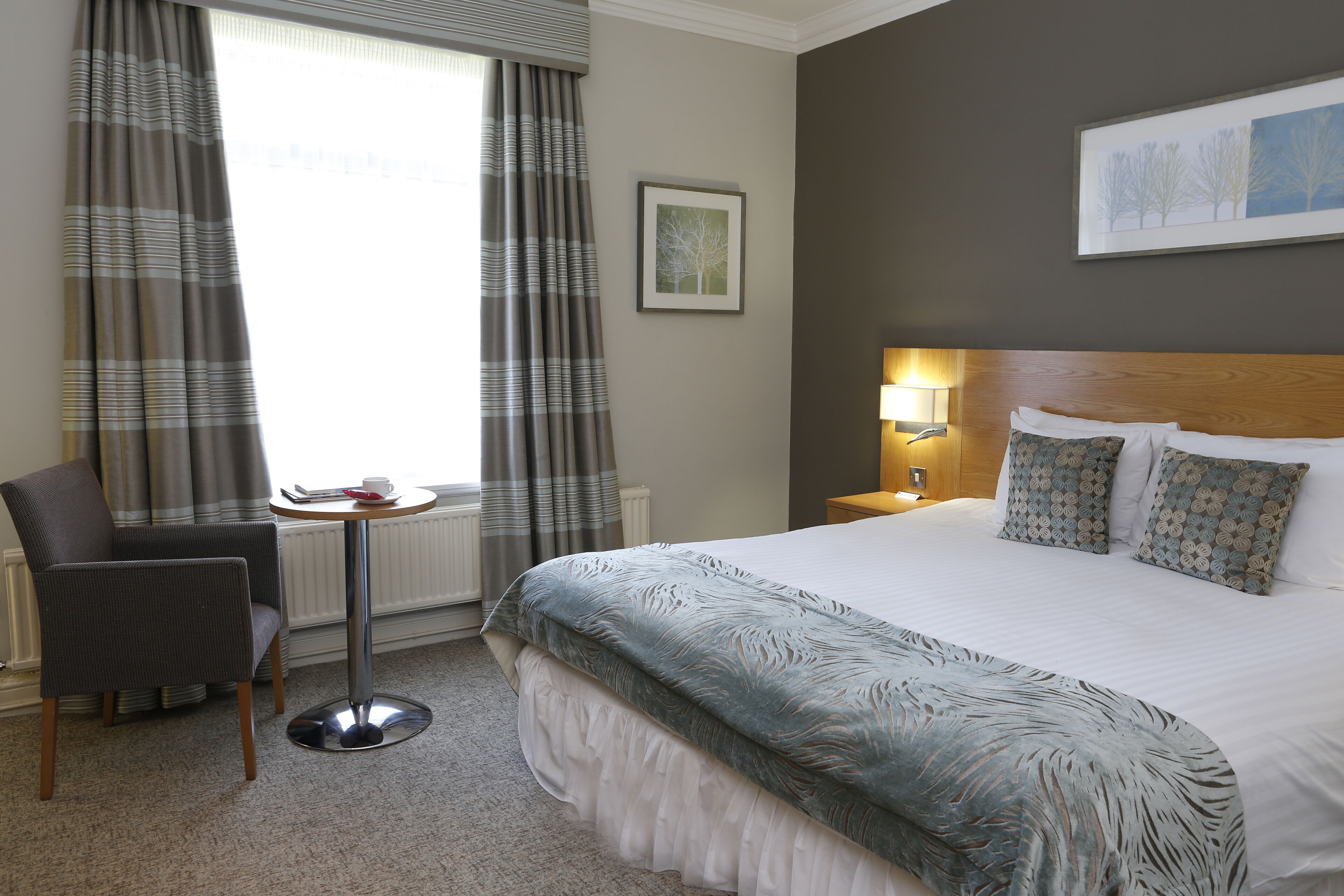 A hotel room with a large bed, a chair, a small table, and a window with curtains.