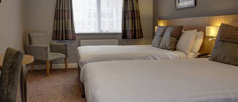 A hotel room with two beds, a chair, a table, a window with curtains, a radiator, and a painting on the wall.