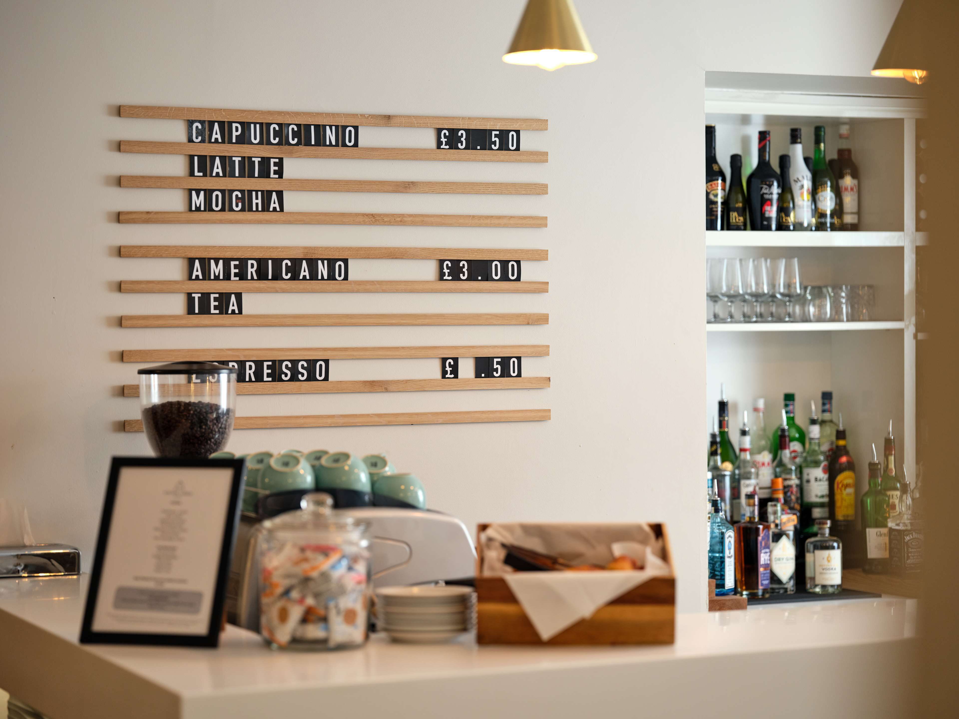 A bar area with a drink menu on the wall listing prices for cappuccino, latte, mocha, americano, tea, and espresso.