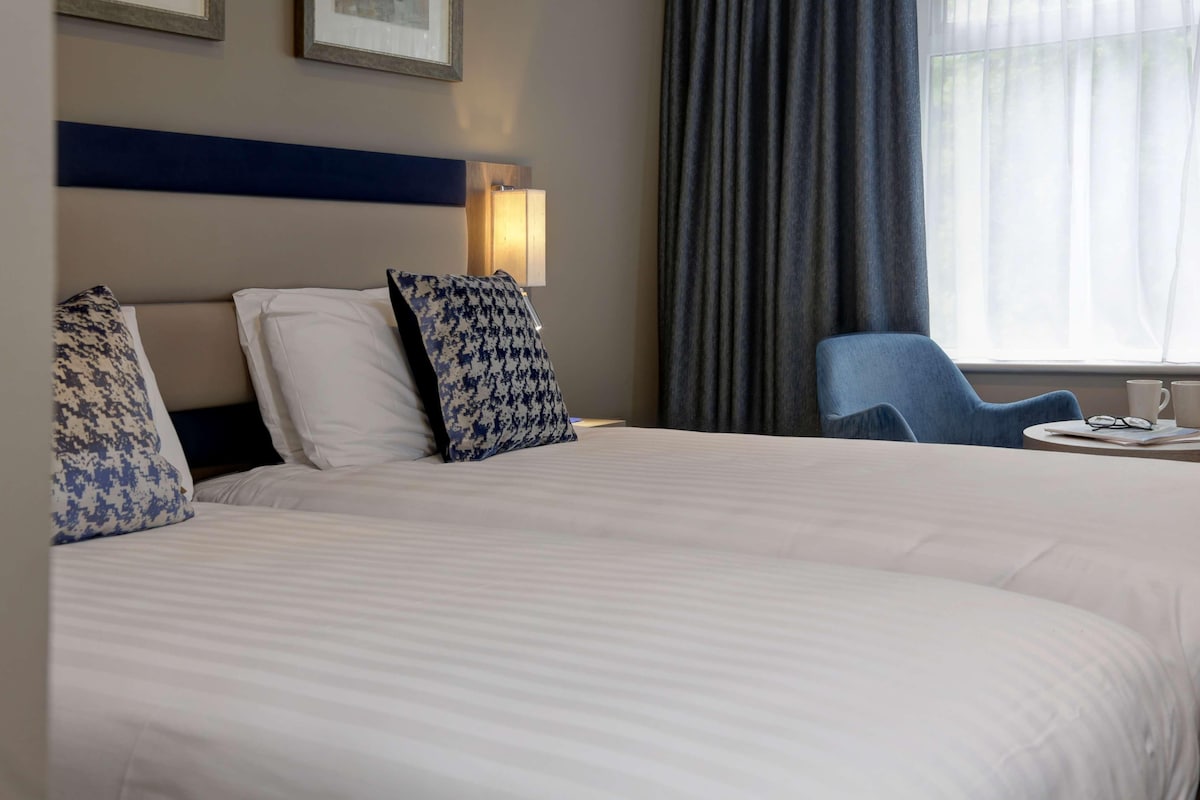 A hotel room with a large bed, two pillows, a bedside lamp, a chair, and a window with curtains.
