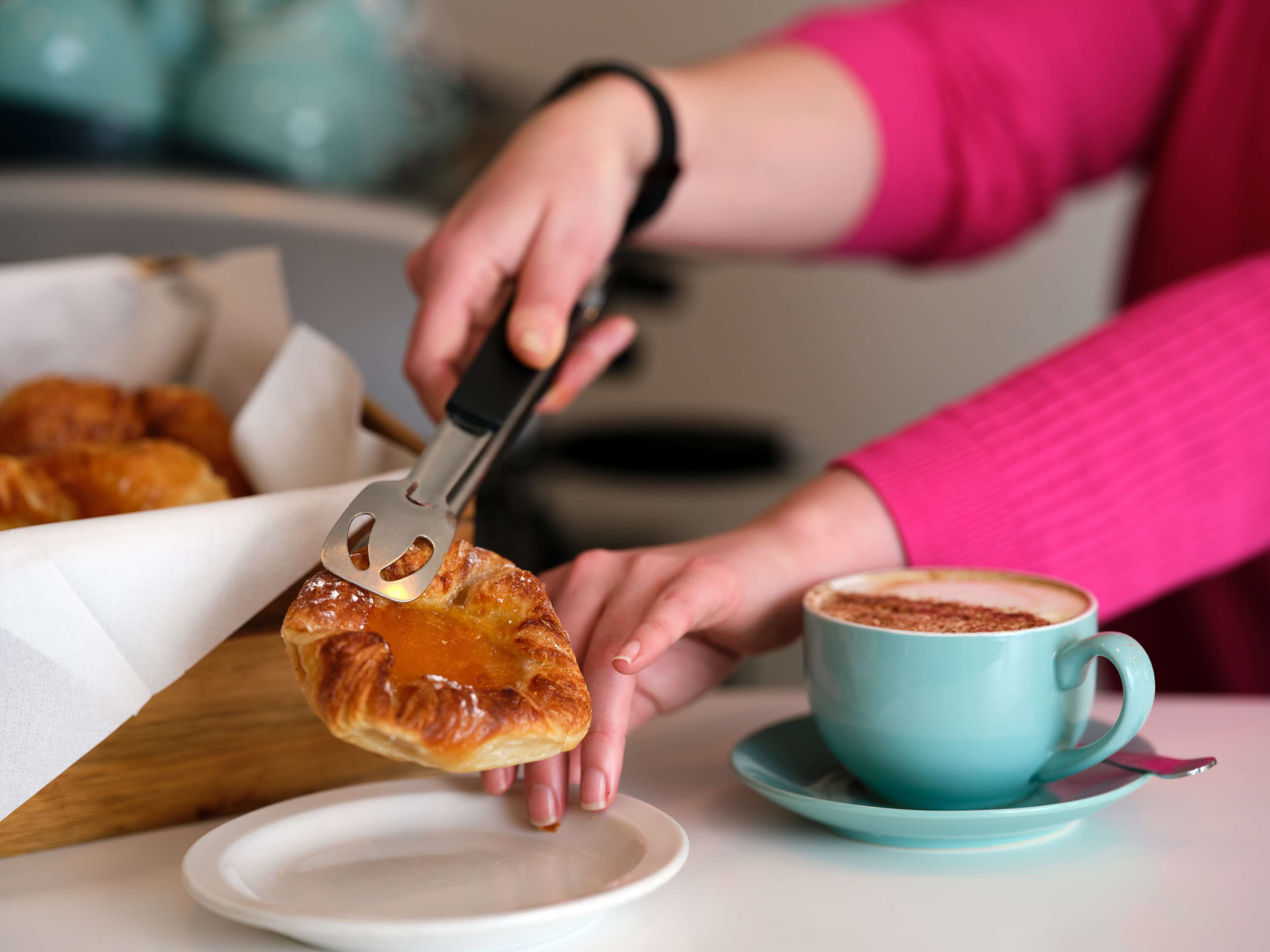 A person holding a pastry with a utensil, a cup of coffee, and a plate on a table.