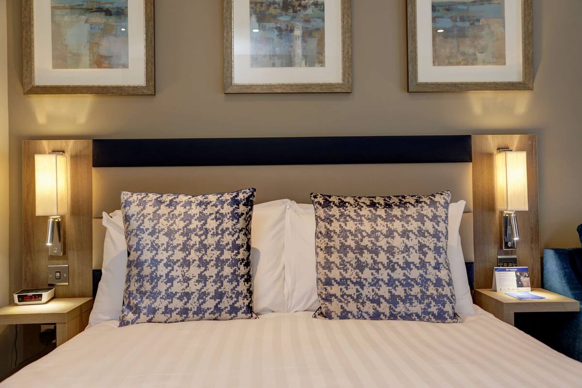 Ein Hotelzimmer mit einem großen Bett, zwei Nachttischen, einer digitalen Uhr, gerahmten Bildern an der Wand und einem blauen Sessel.