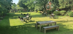A well-maintained backyard with a wooden table and chairs, surrounded by lush greenery and a brick building in the background.