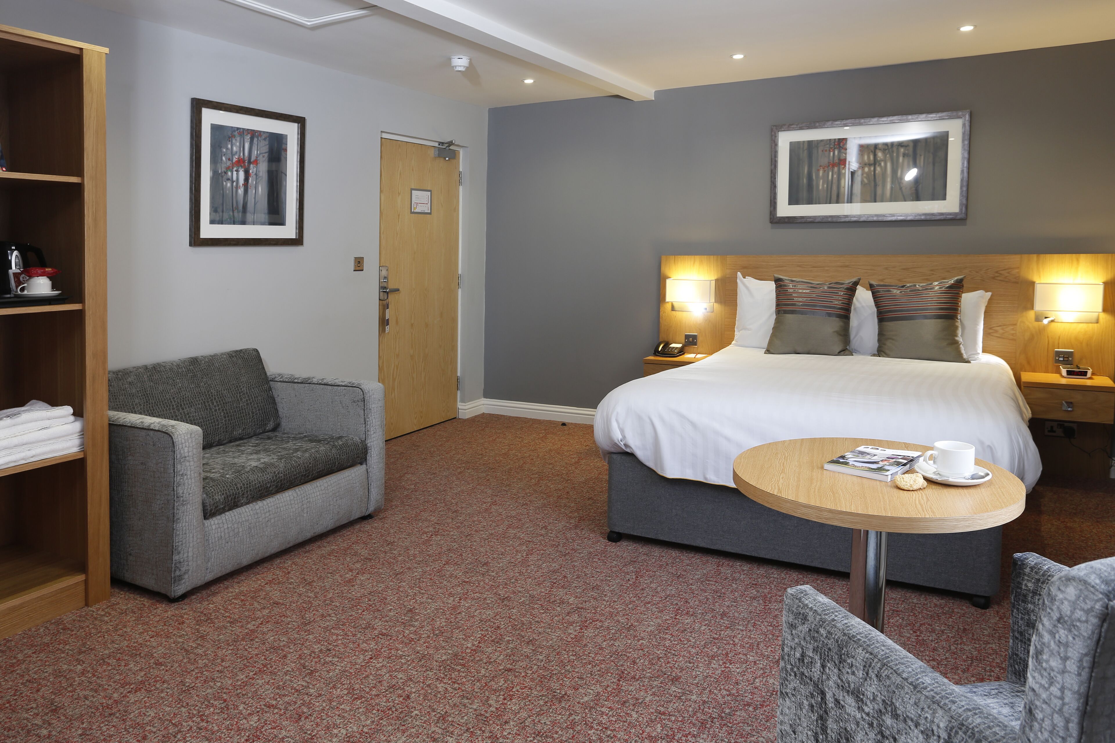 A hotel room with a bed, two bedside tables, a round table with a cup and a book, a grey armchair, and a bookshelf.