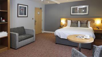Une chambre d’hôtel avec un lit, deux tables de chevet, une table ronde avec une tasse et un livre, un fauteuil gris et une bibliothèque.