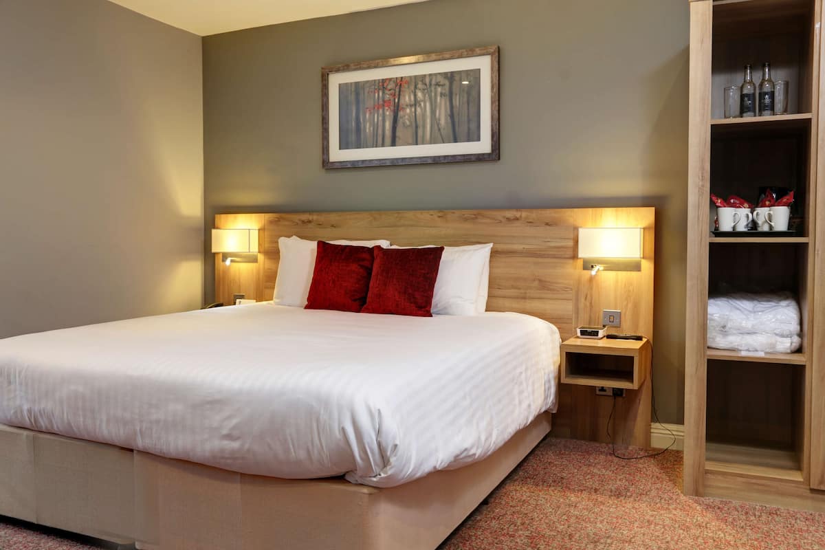 Ein Hotelzimmer mit einem großen Bett, zwei Nachttischen mit Lampen, einem Holzkopfende, einem gerahmten Bild an der Wand und einem Schrank mit Flaschen und Gläsern.