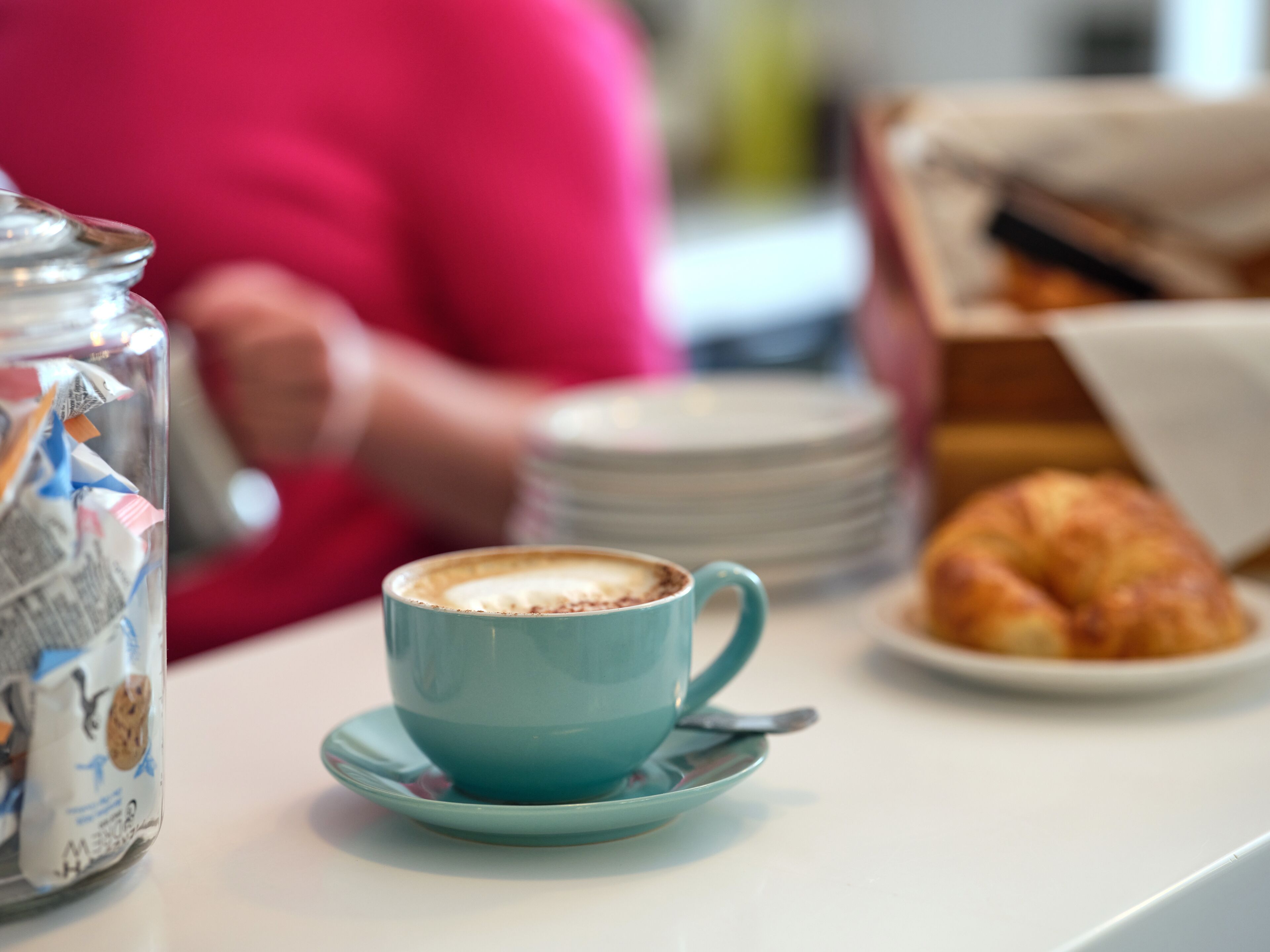 A table with a blue cup of coffee, a croissant, and a jar of cookies.