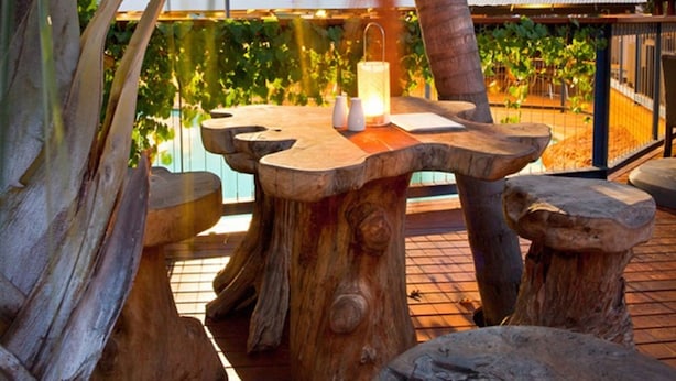 Un área de estar al aire libre con madera, con una mesa y bancos, rodeada de palmeras y un viñedo al fondo.