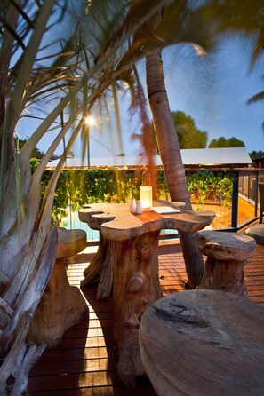 Un espace extérieur aménagé avec des meubles en bois, comprenant une table et des bancs, entouré de palmiers et avec un vignoble en arrière-plan.