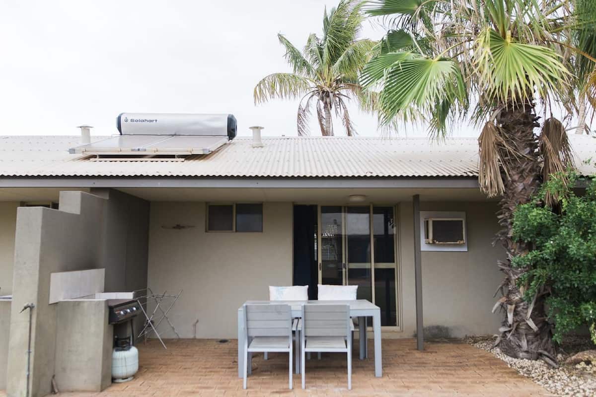 Une maison avec un réservoir d’eau sur le toit, une terrasse avec une table et des chaises, et un palmier.