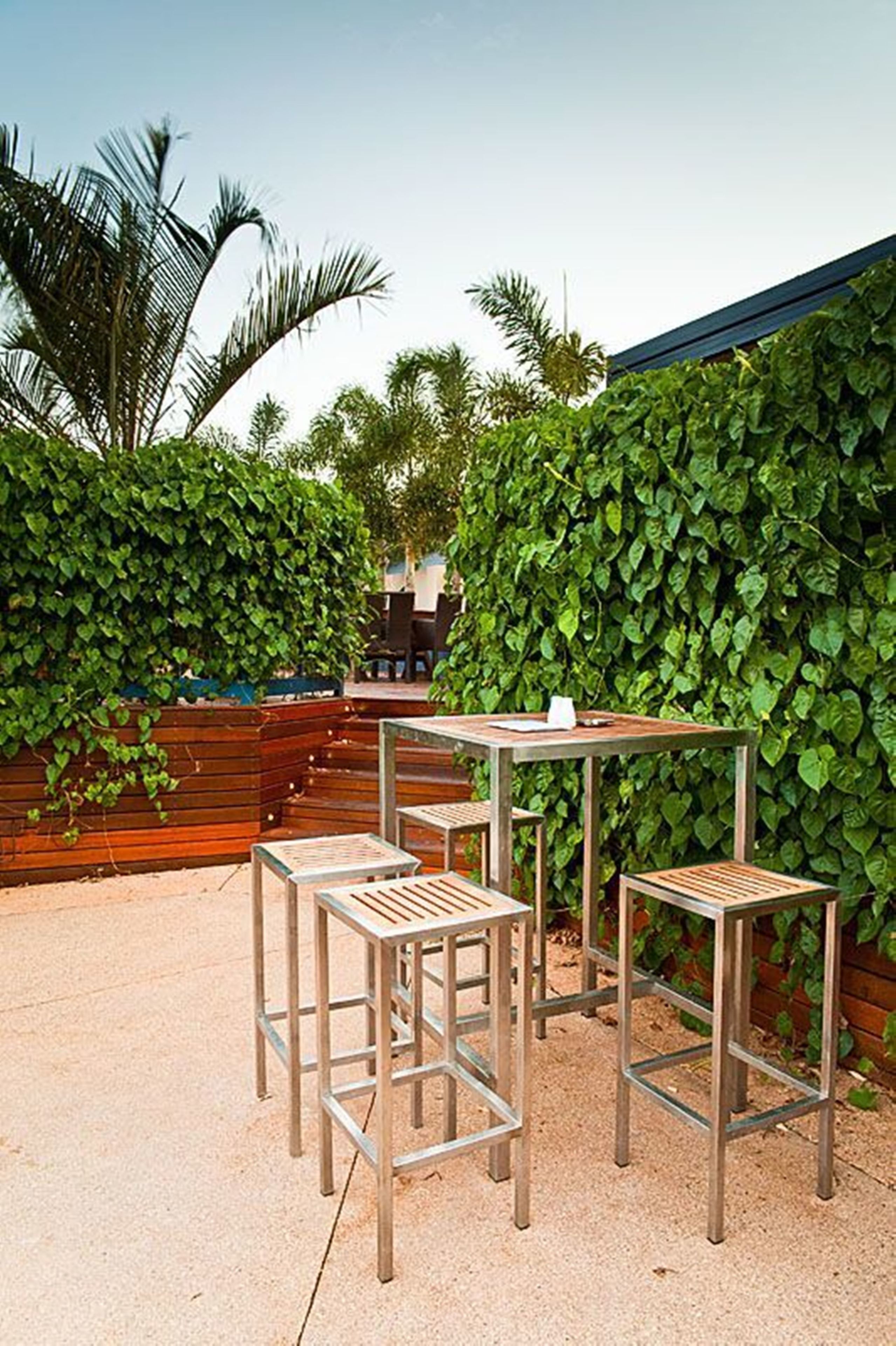 Een terras met metalen tafels en stoelen, omgeven door een weelderige groene haag.