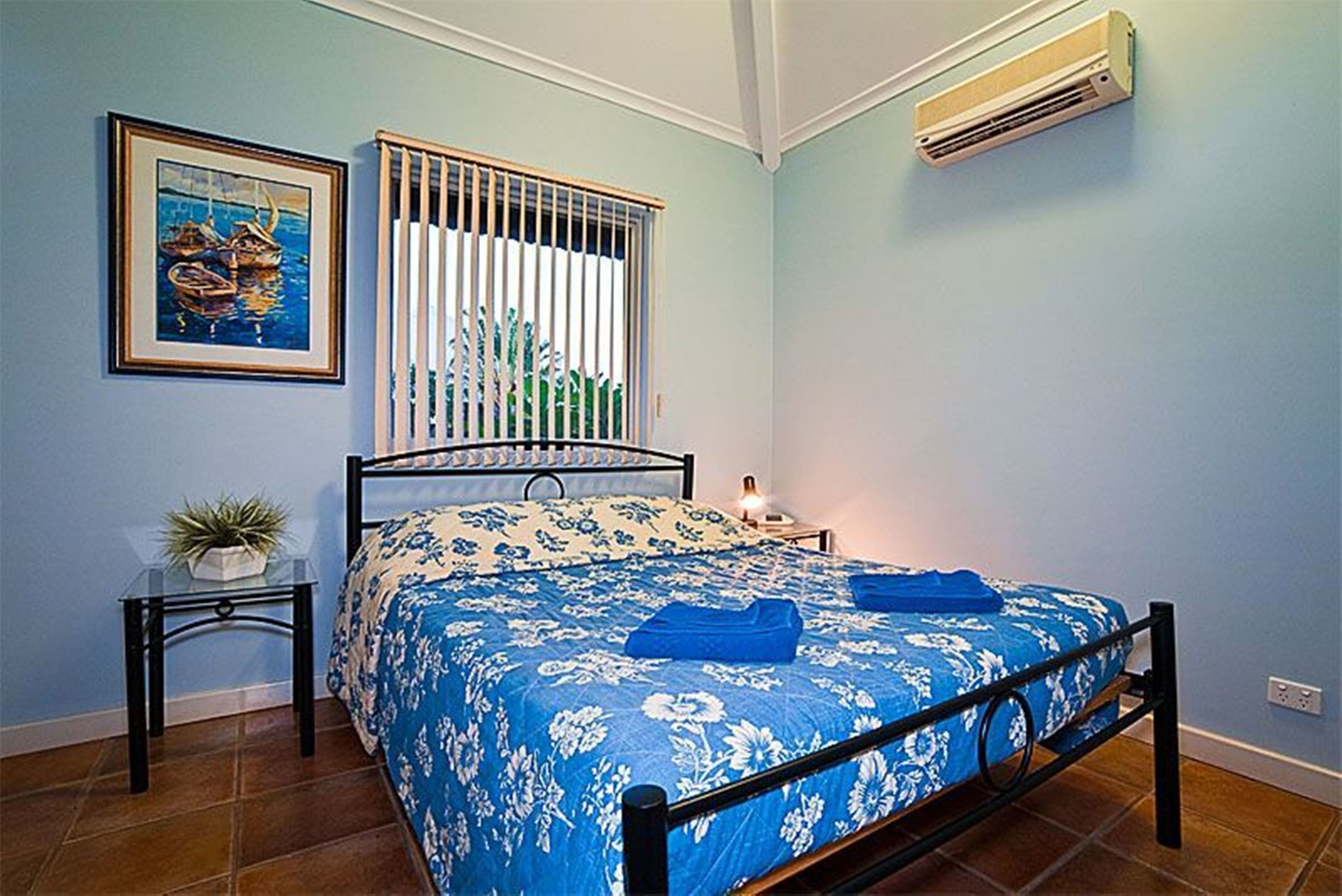 Un dormitorio con cama, mesita de noche, planta en maceta, ventana con persianas y equipo de aire acondicionado.