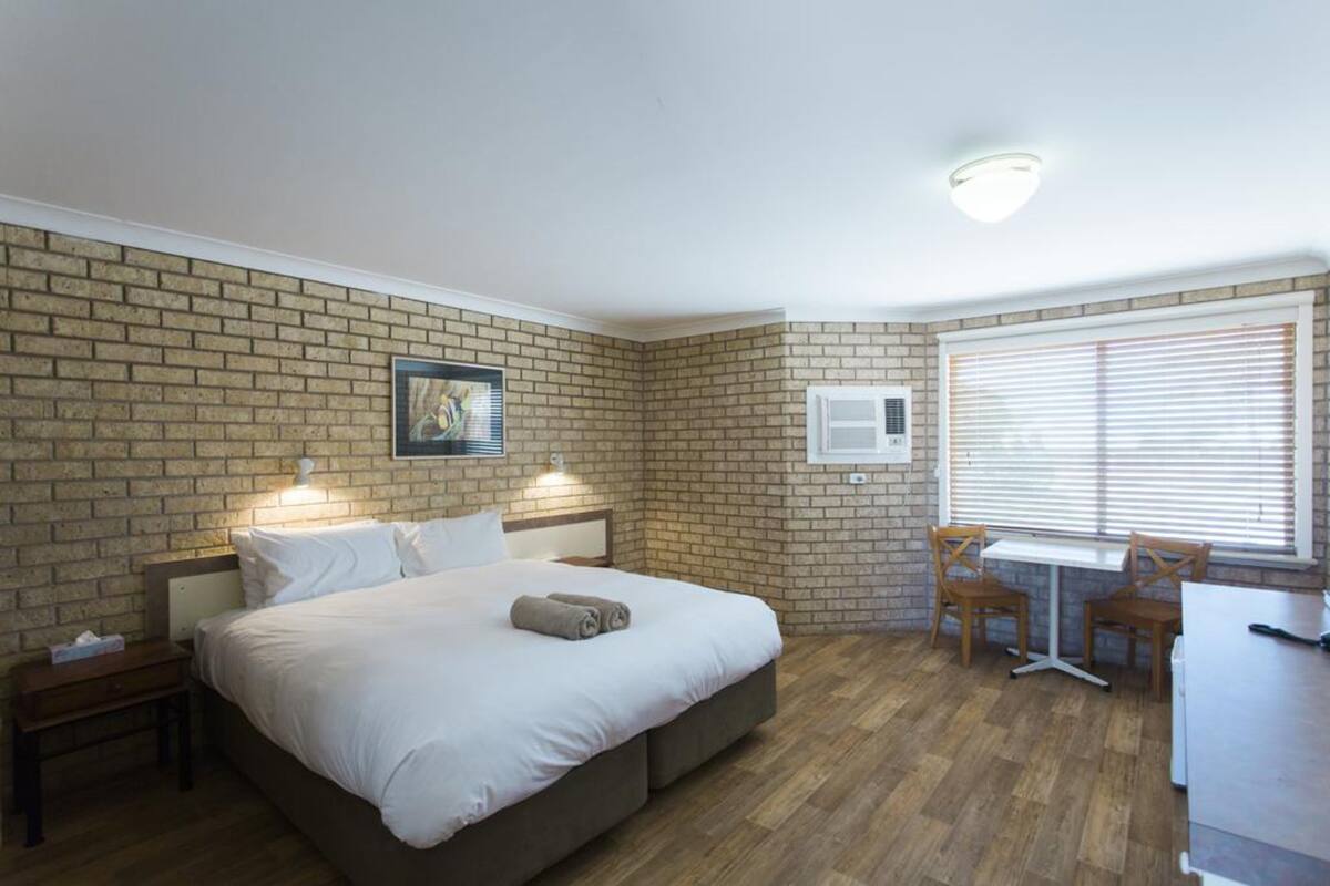 Une chambre d’hôtel avec un mur en briques, un lit avec du linge de maison blanc, une petite table avec des chaises et une fenêtre avec des stores.