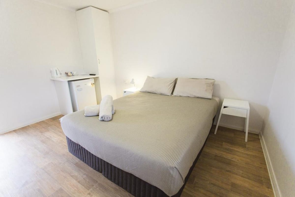 Une chambre d’hôtel bien rangée, avec un lit, une petite table de chevet, une commode et un bureau avec une chaise.
