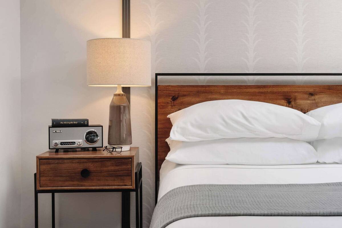 Een netjes opgemaakt bed met witte kussens en een grijze deken, een bedlampje en een radio op een nachtkastje van hout.