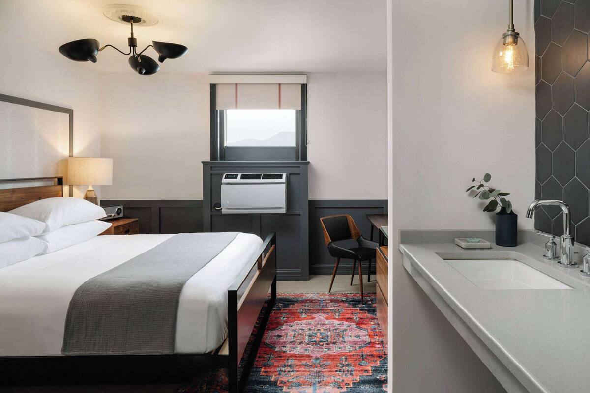 Hotelkamer met een bed, nachtkastje, lamp, bureau, stoel, raam, airconditioning en een badkamer met een wastafel en spiegel.