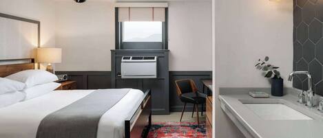 A hotel room with a bed, bedside table, lamp, desk, chair, window, air conditioning unit, and a bathroom with a sink and mirror.
