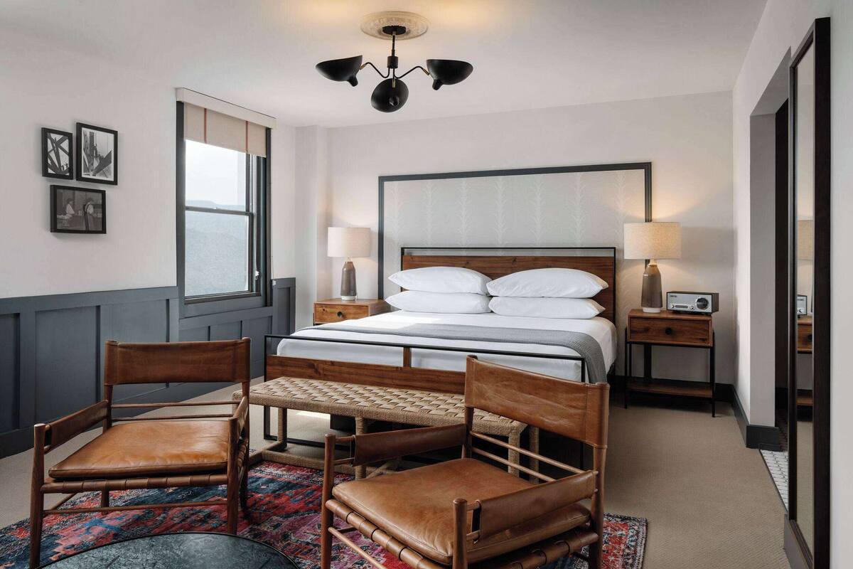 Een hotelkamer met een bed, twee nachtkastjes, een bureau met stoelen, een raam en een plafondlamp.