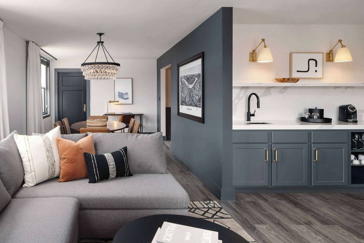 Een moderne woonkamer met een grijze bank, een kroonluchter en een keuken met grijze kasten en een wit aanrecht.