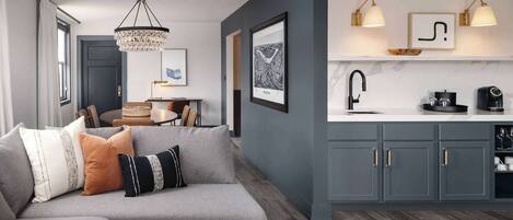 A modern living room with a grey sofa, a chandelier, and a kitchen area with grey cabinets and a white countertop.