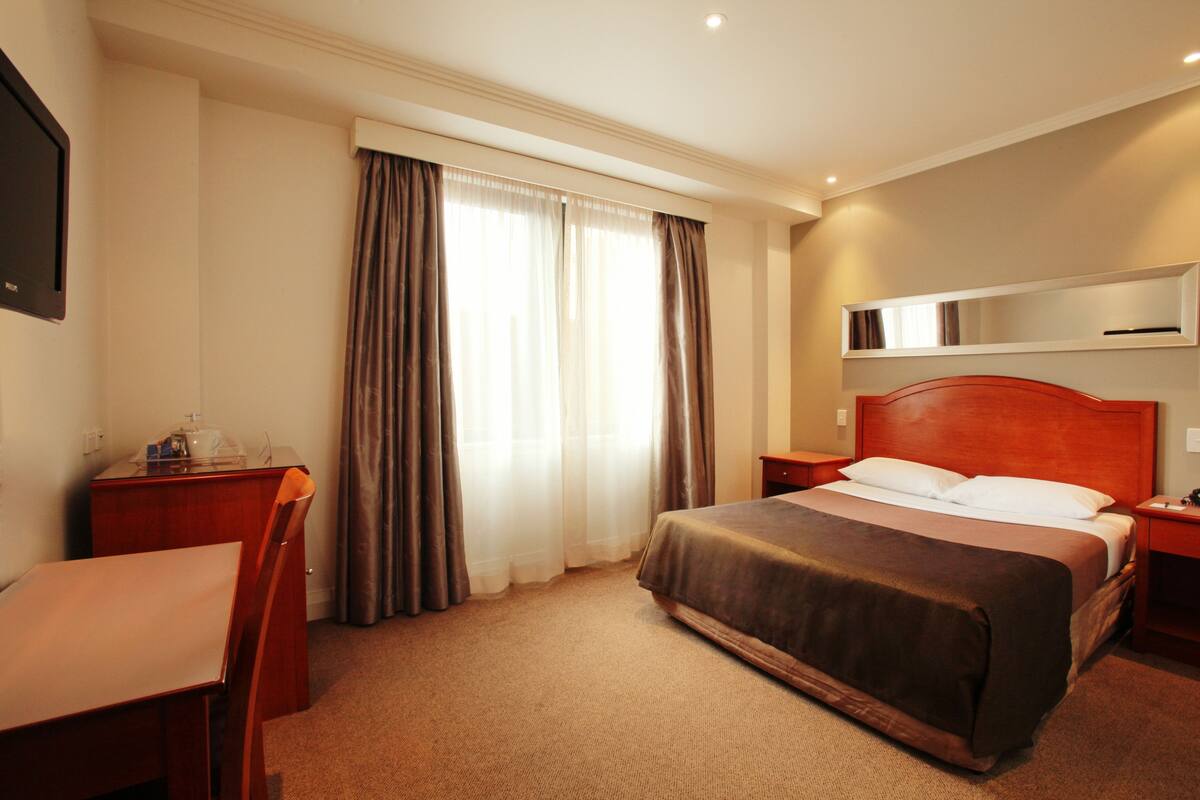 Habitación de hotel con cama, escritorio, silla, mesita de noche y ventana con cortinas.