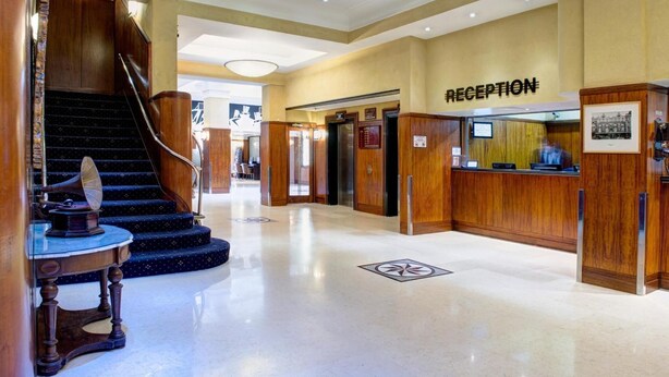 Zona de recepción de un hotel con un mostrador de madera, un cartel de recepción y una escalera que lleva a un piso superior.