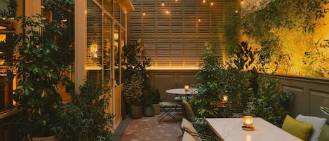 An indoor patio area with tables and chairs, surrounded by potted plants and a tiled floor.