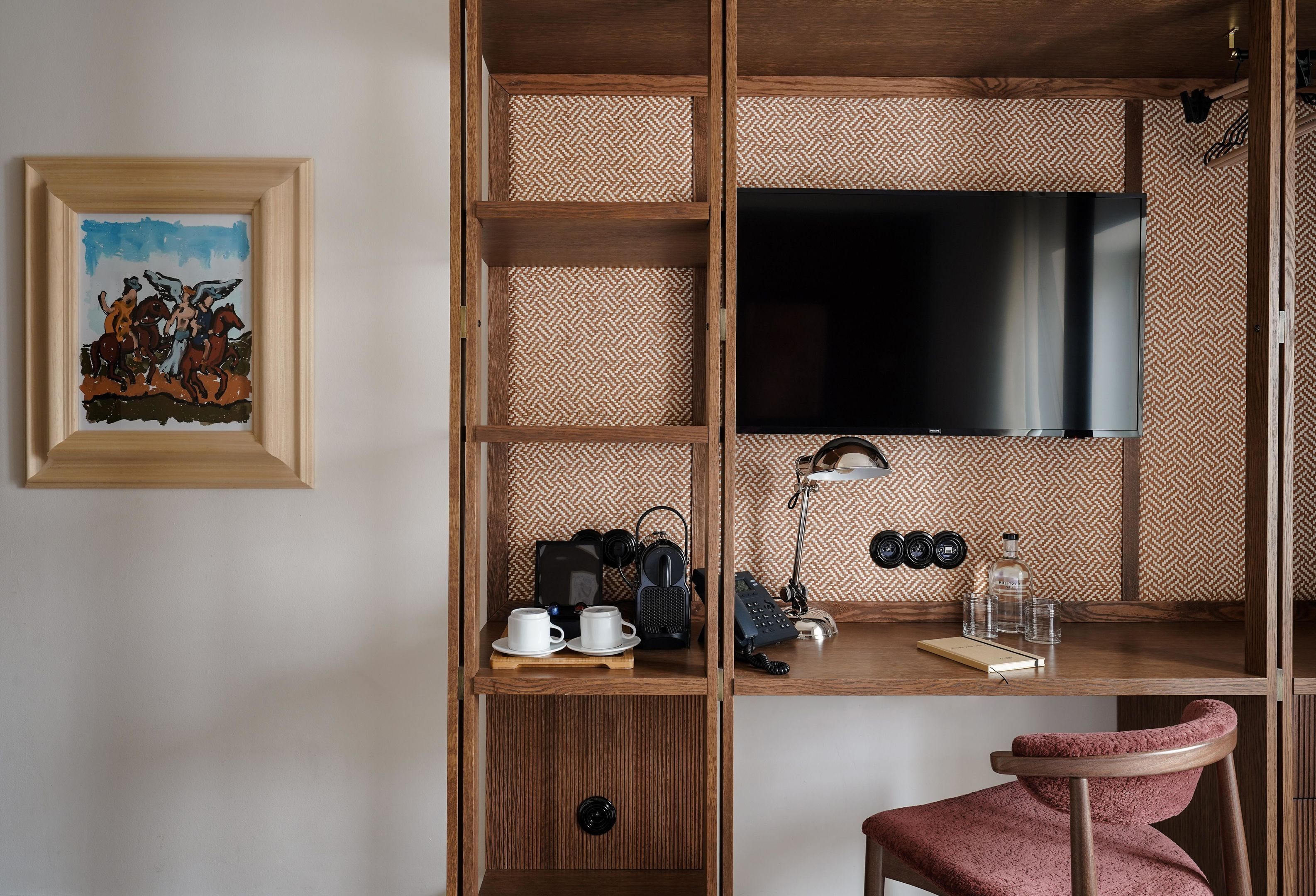 Une pièce de taille modeste, avec un bureau en bois, un téléviseur fixé au mur, un tableau encadré et une chaise rouge.