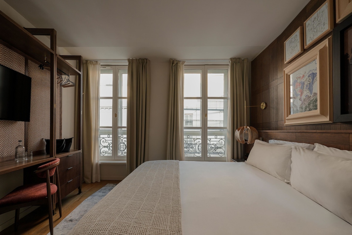 A hotel room with a bed, a desk, a television, and large windows with curtains.