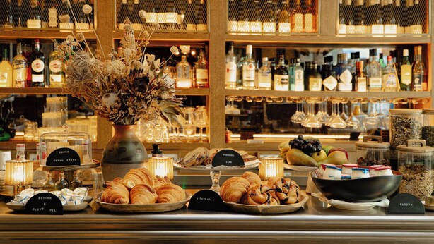 A bar area with various bottles and glasses, a vase with dried flowers, and labeled food items like croissants and pastries. - Hotel Pulitzer Paris (Paris)