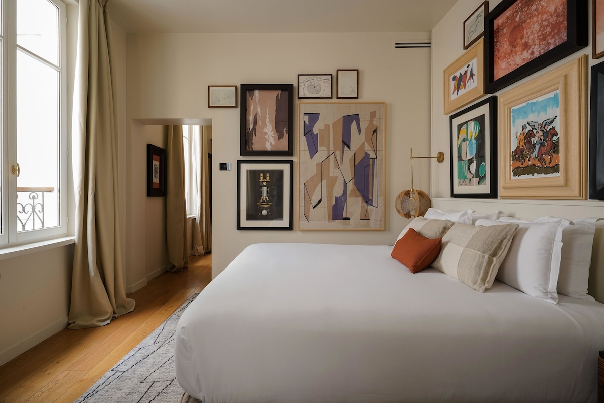 A bedroom with a large bed, multiple framed artworks on the wall, and a window with curtains.