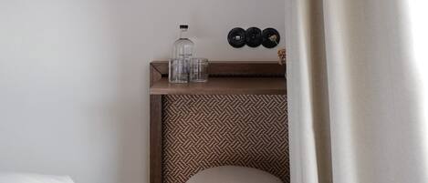 A hotel room with a bed, a small wooden side table with a bottle and glasses, a framed picture on the wall, and a patterned rug.