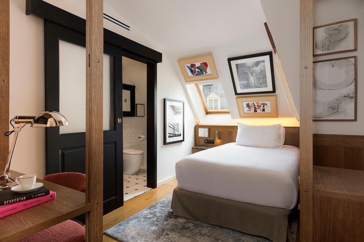 A hotel room with a bed, a desk with a lamp, a chair, and a bookshelf. There are framed pictures on the wall and a window with a view of the outside.